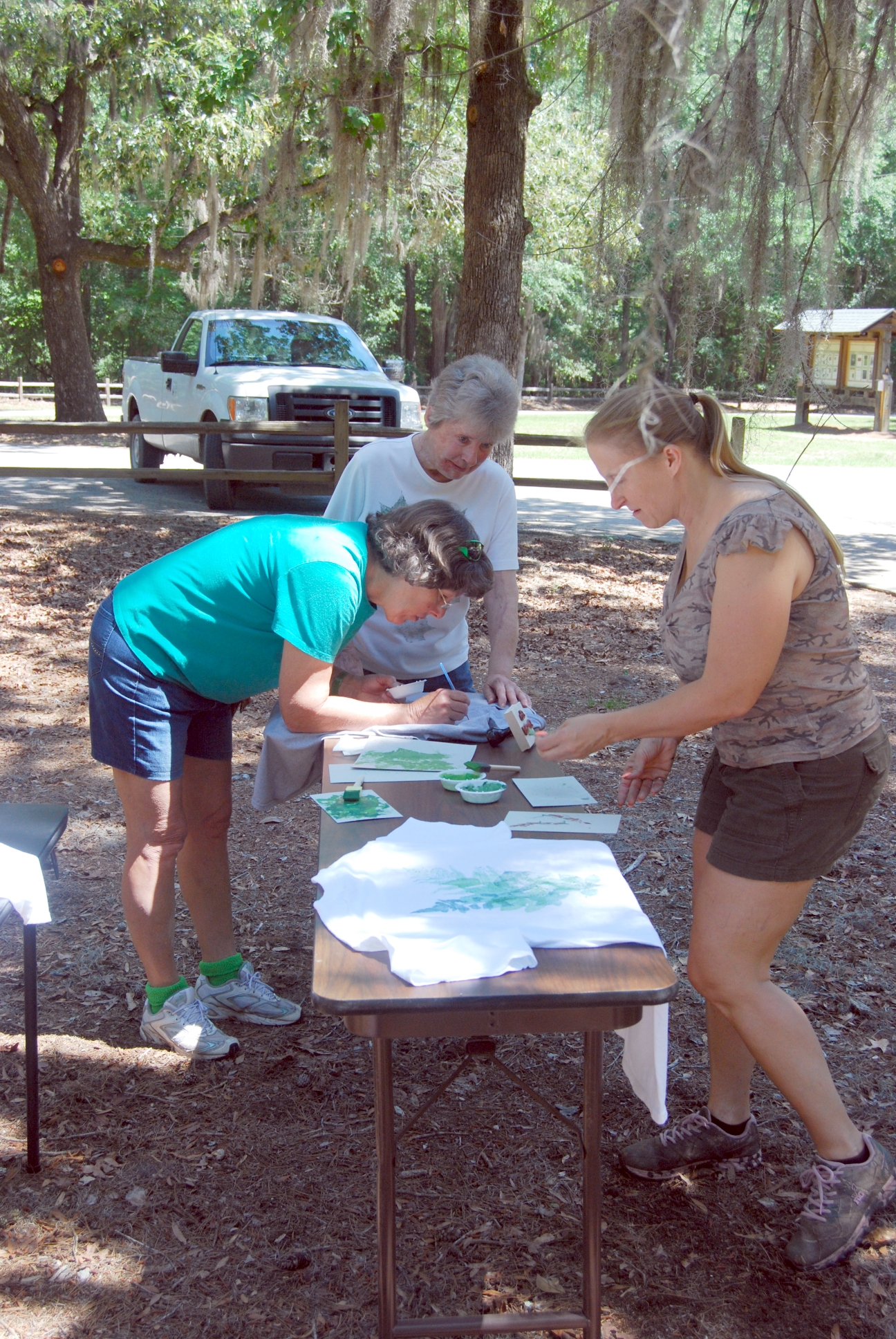 Three woman make art at a table set up in a wooded area.
