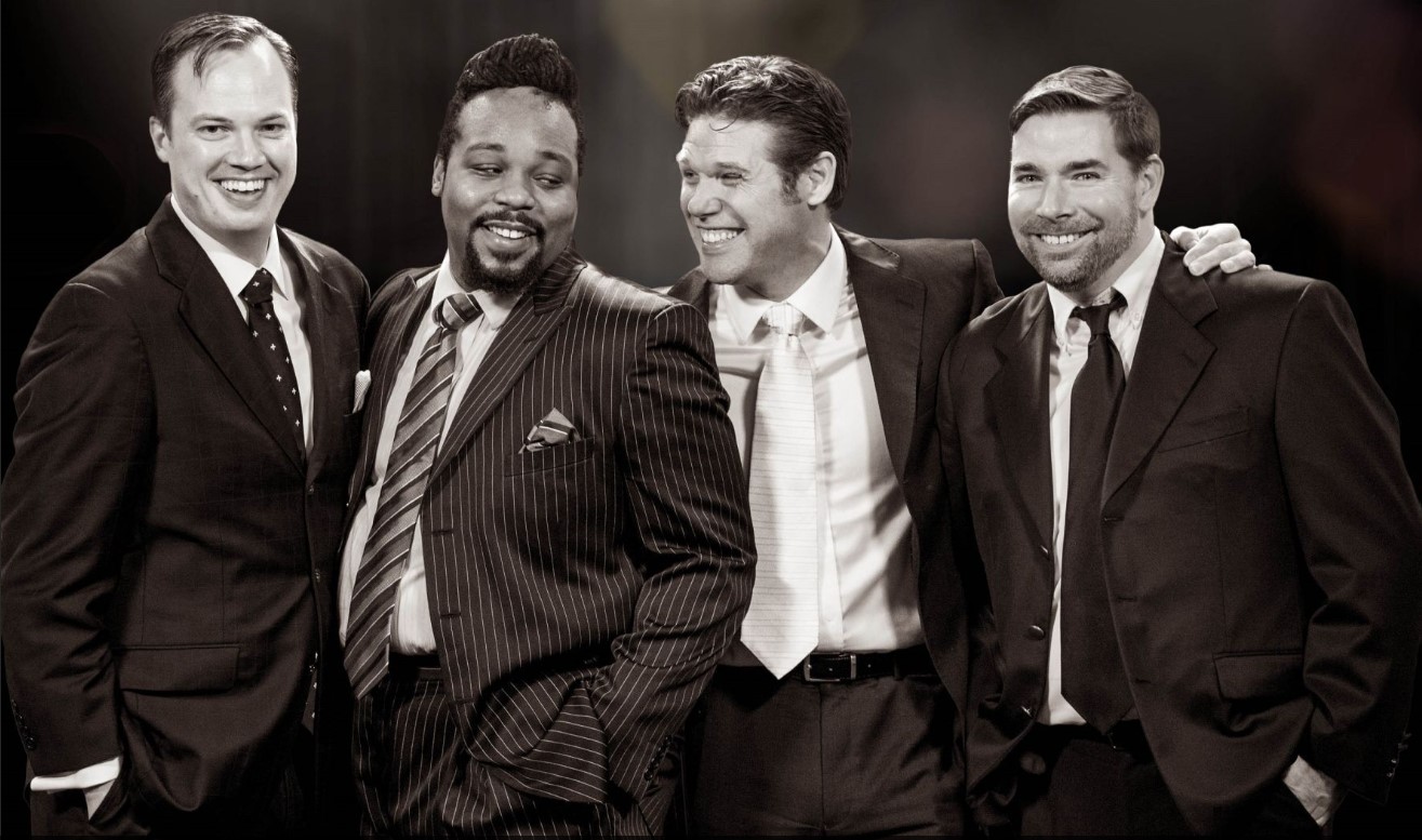 Four Rat Pack-styled men, one Black, pose laughing with arms around each other.