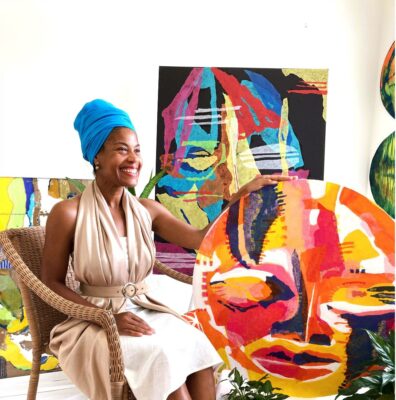 Person presenting as BIPOC female sits, smiling, with her brightly colored graphic artwork.