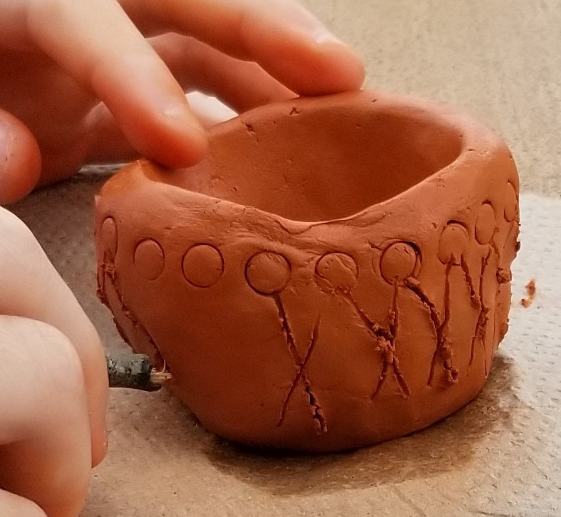 Hands etching a small clay pot being formed.