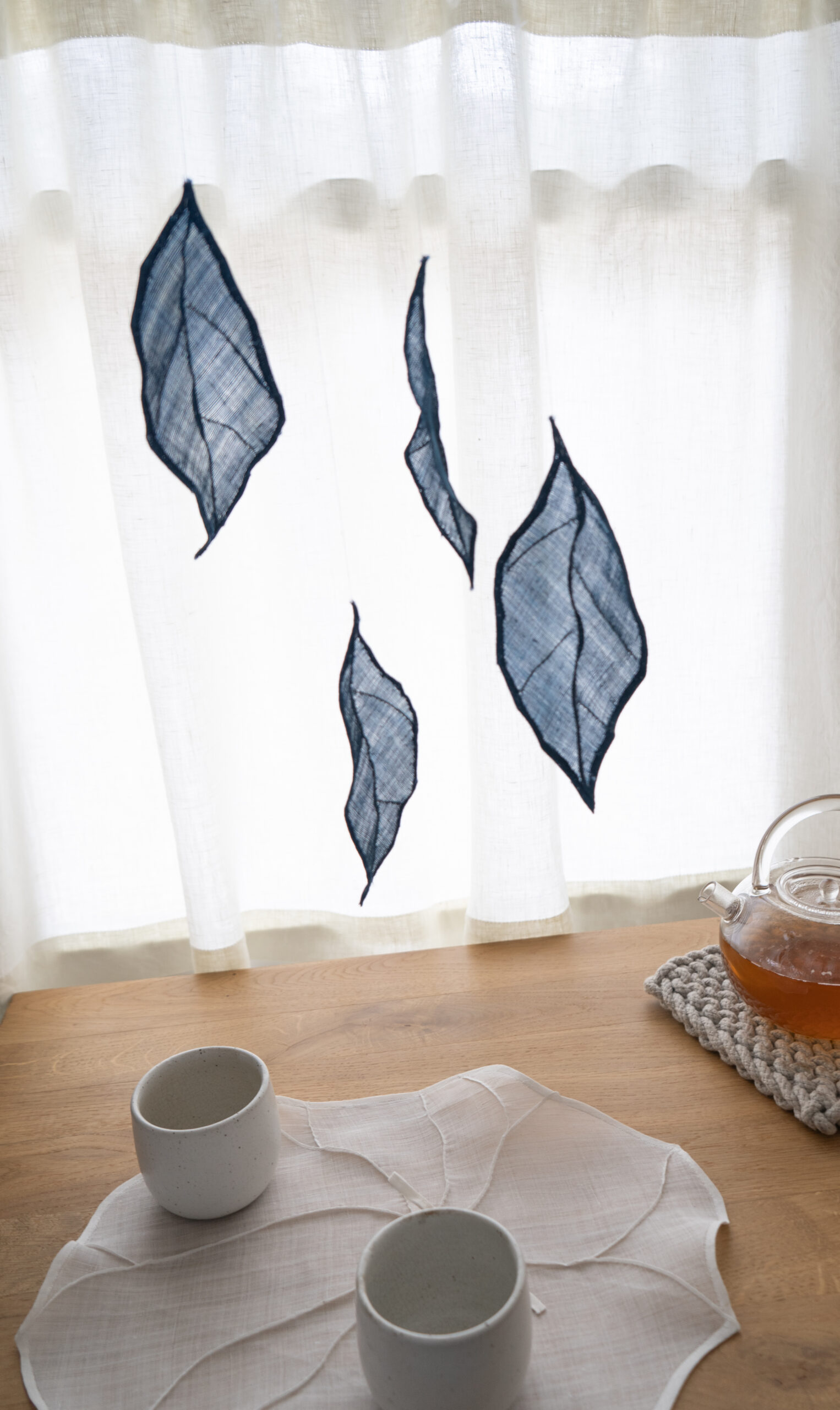A mobile of blue translucent fabric leaves, over a white fabric leaf shape and white ceramic cups.