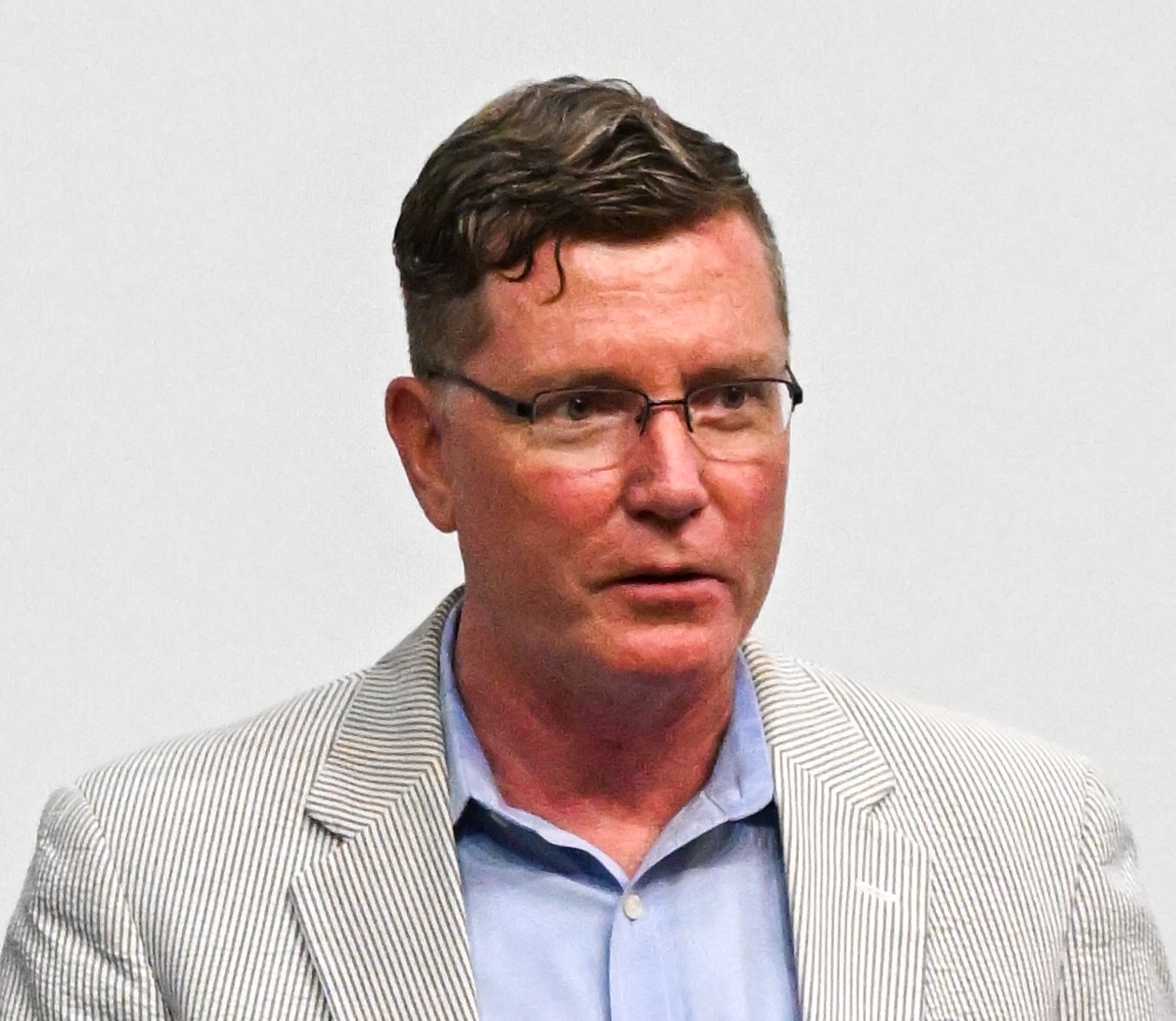 A man with short dark hair and glasses, wearing a seersucker jacket and blue button-front shirt.