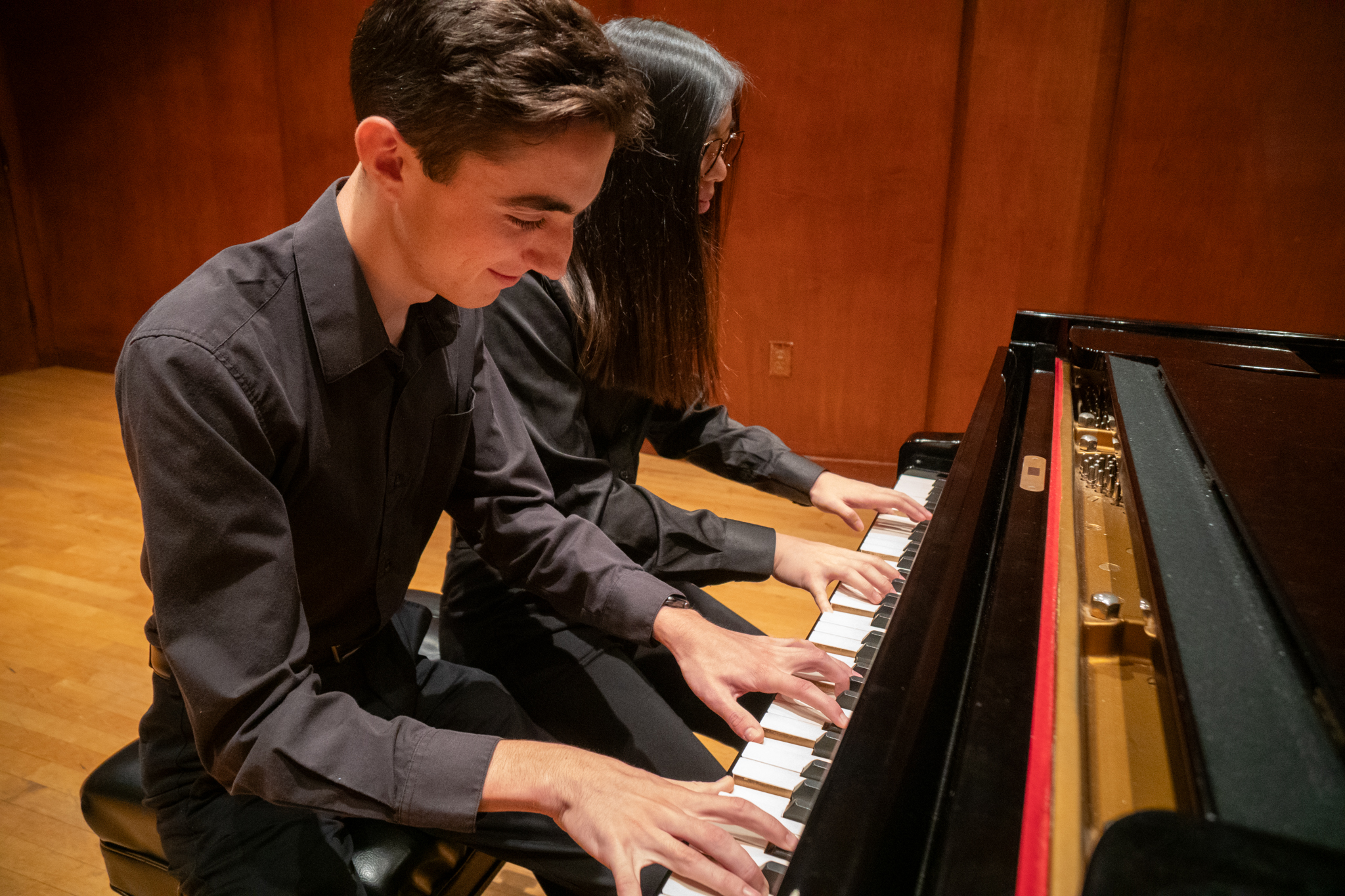 Two students play a four-handed piece on a grand piano.