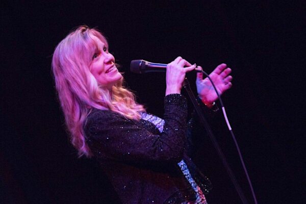 A blonde woman in a black sequined jacket sings at an onstage microphone.