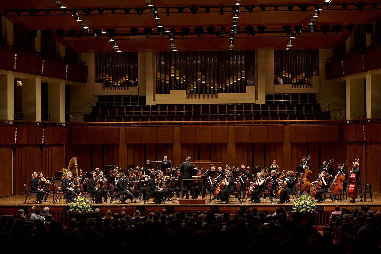 An orchestra performing on a stage in a large concert hall, in front of a large pipe organ.