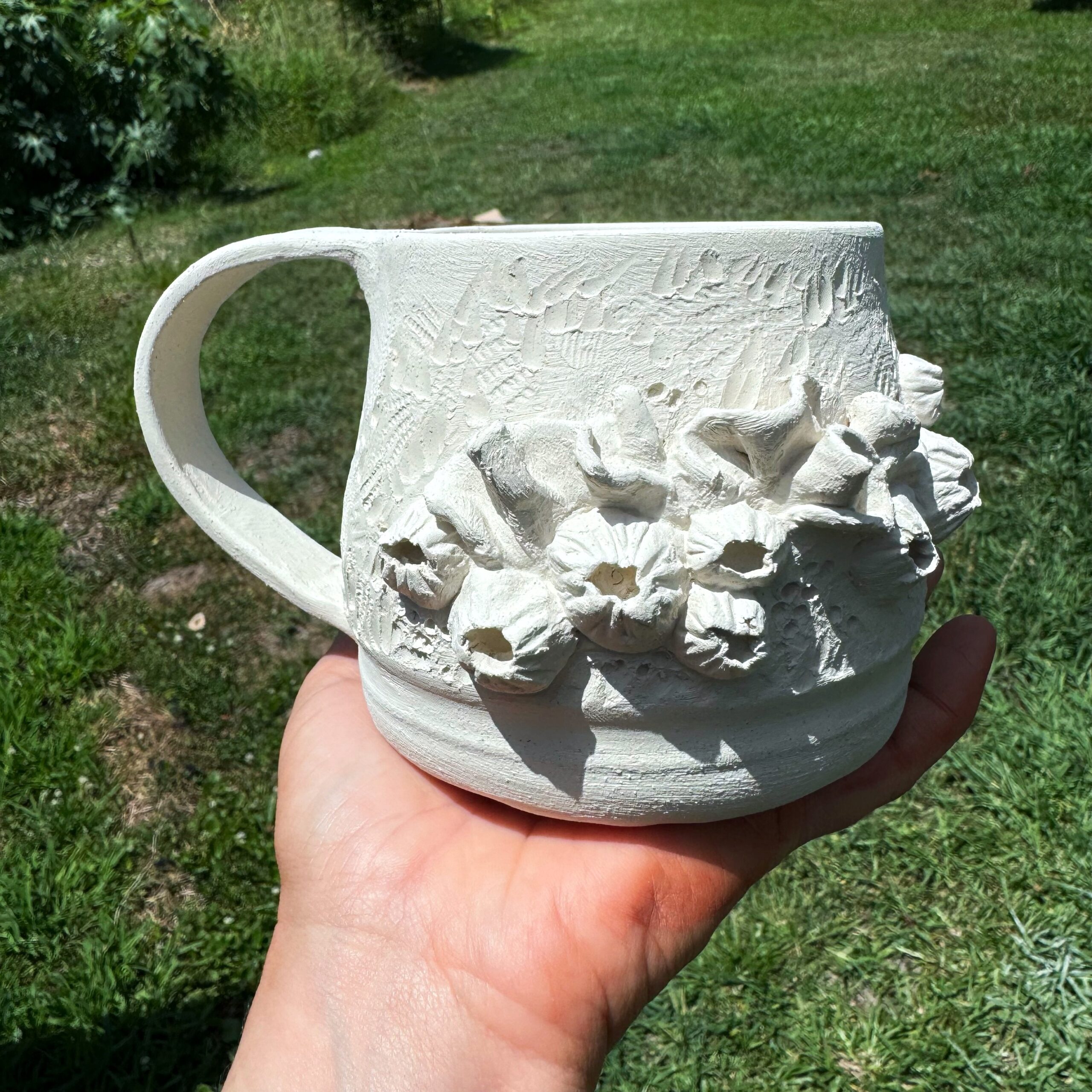 A hand holds a hand-built ceramic mug outdoors. The mug is white with 3-dimensional flowers.