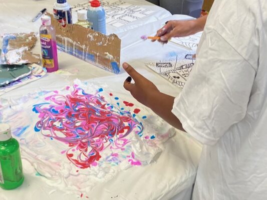 A person in a t-shirt stands over swirls of paint on a flat work surface.