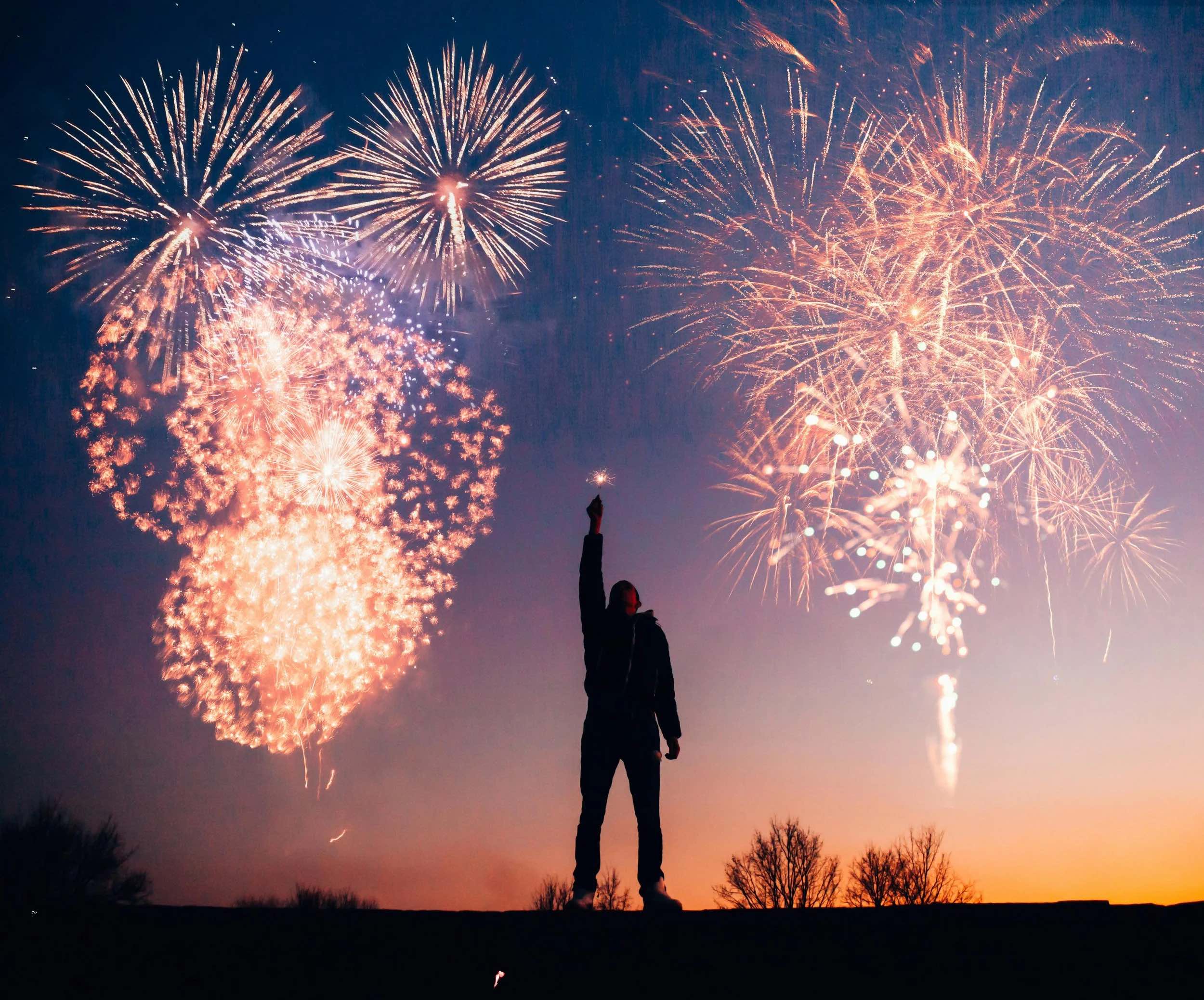 A man stands on a ridge holding up a lit sparkler, as fireworks explode in the sky behind him.