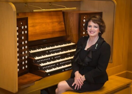 A smiling woman in black formalwear sits at an organ with 4 keyboards and 2 stop jams.