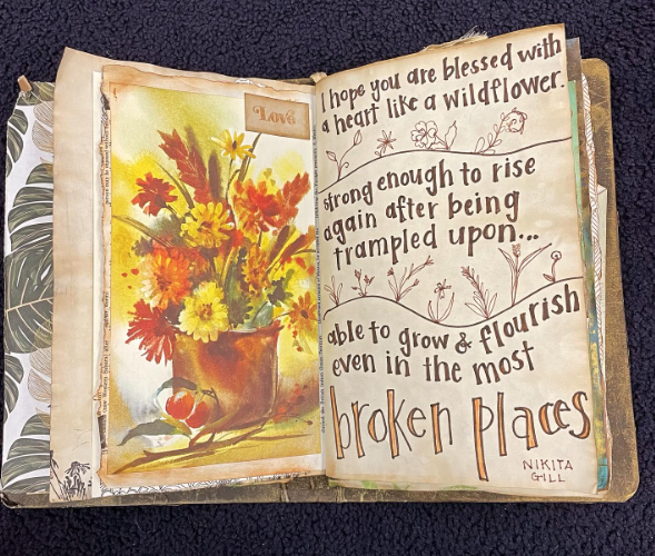 An artwork book is open to a painting of autumnal wildflowers in a brown pot.