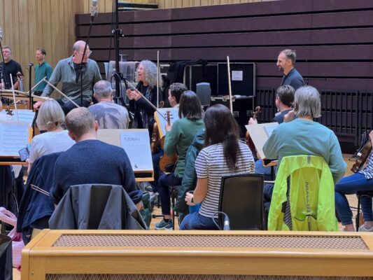 A white woman with wavy gray hair stands amidst an orchestra in casual clothing.