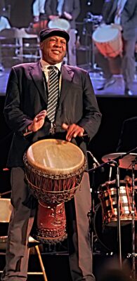 A Black man in a coat, tie, and cap plays an African drum onstage with other musicians.