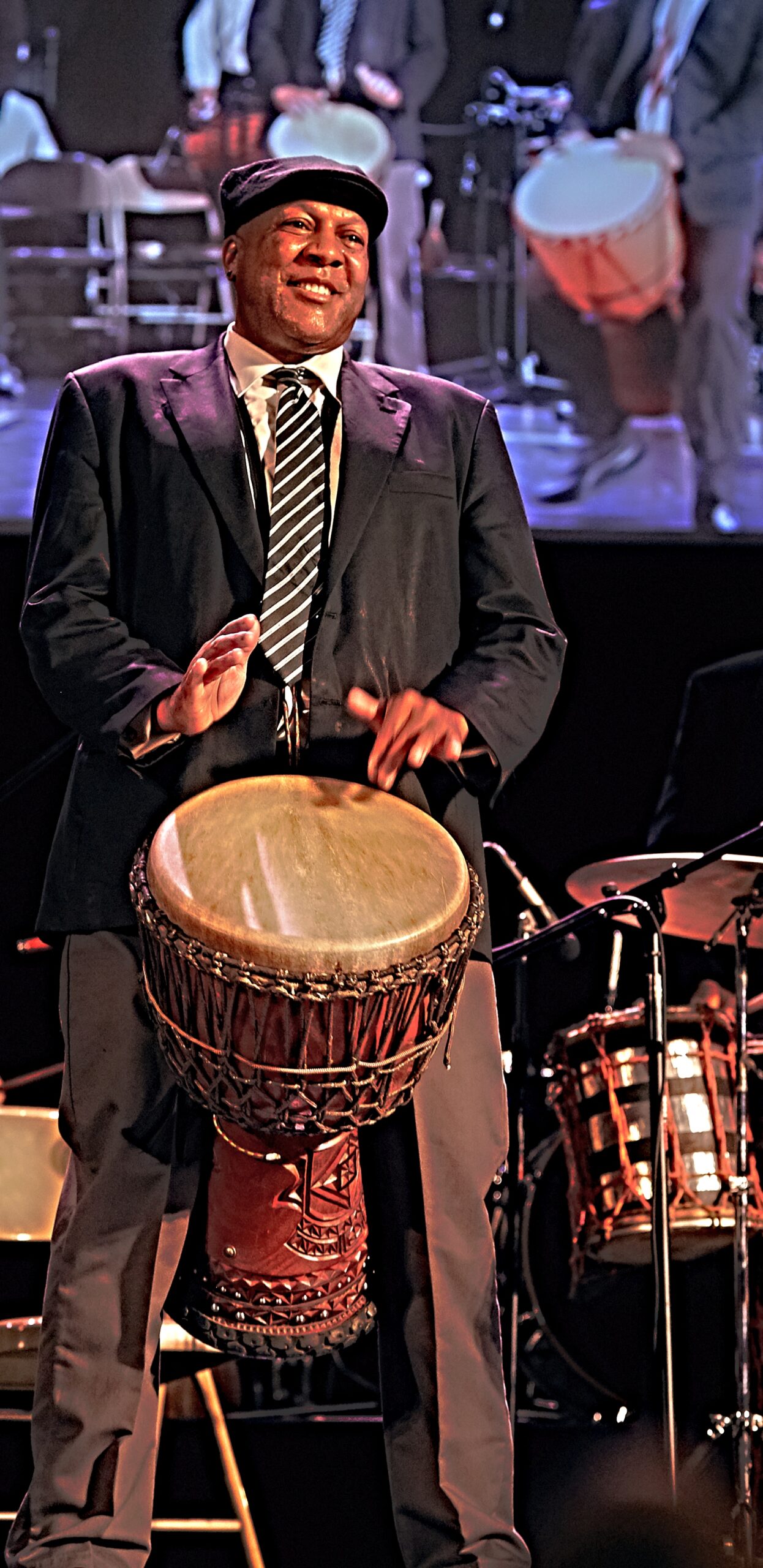 A Black man in a coat, tie, and cap plays an African drum onstage with other musicians.