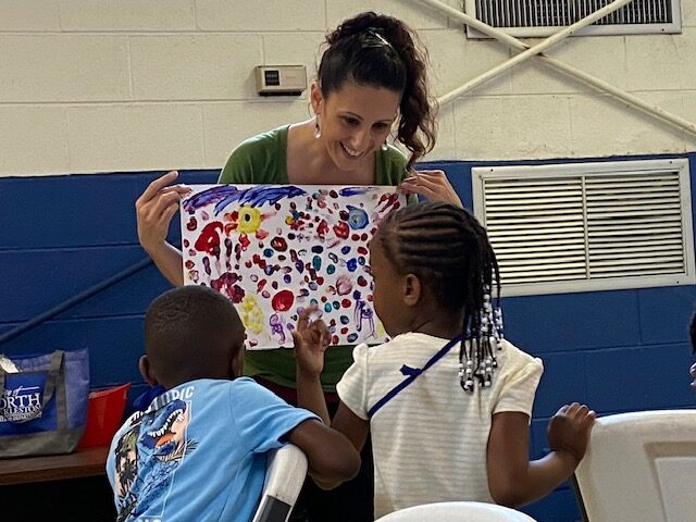 A smiling woman holds up a colorful piece of art for two Black children to see.