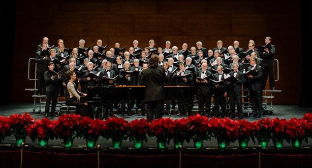 A men's choir in tuxedos sings on a stage decorated with poinsettias.