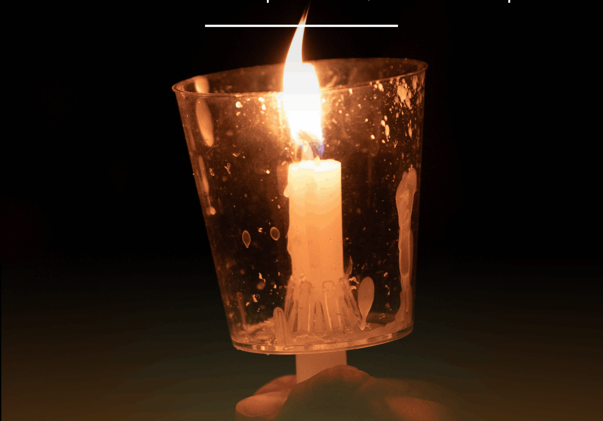 A white candle burns in a clear cup, held in a hand.