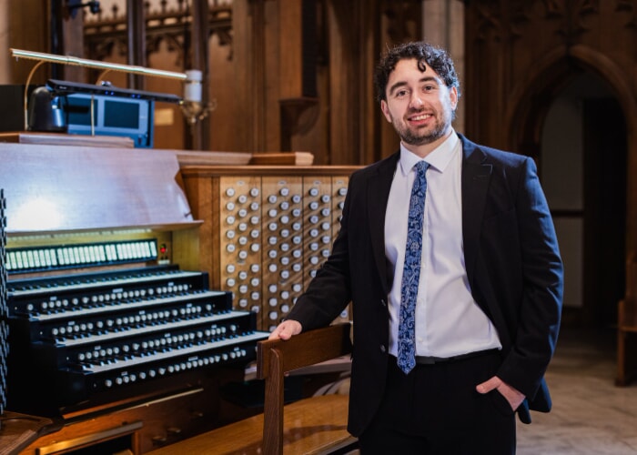 A man with dark curly hair and facial hair smiles by a pipe organ.