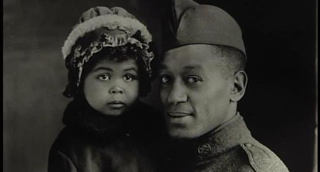 A Black United States soldier from World War I holds a young child in a winter coat and hat.