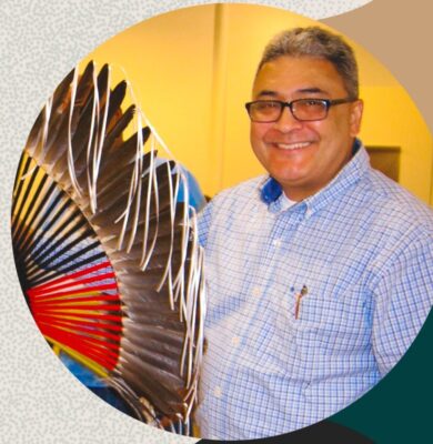 A smiling Native American man with glasses holds a Native American object with feathers.