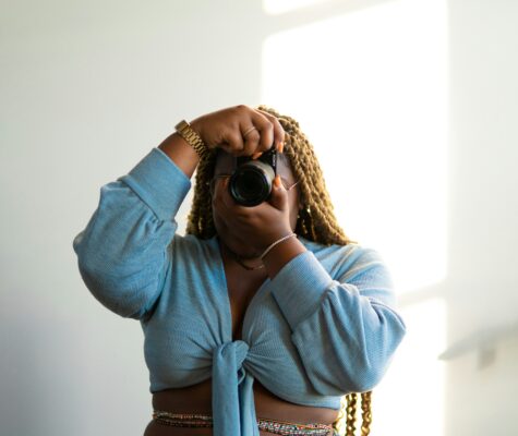 A Black woman holds a DSLR camera to her eyes, pointing toward the viewer.
