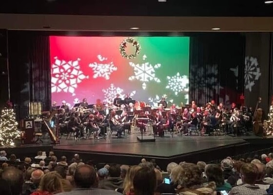 A concert band on stage with Christmas and winter decorations. An audience is seated in front.