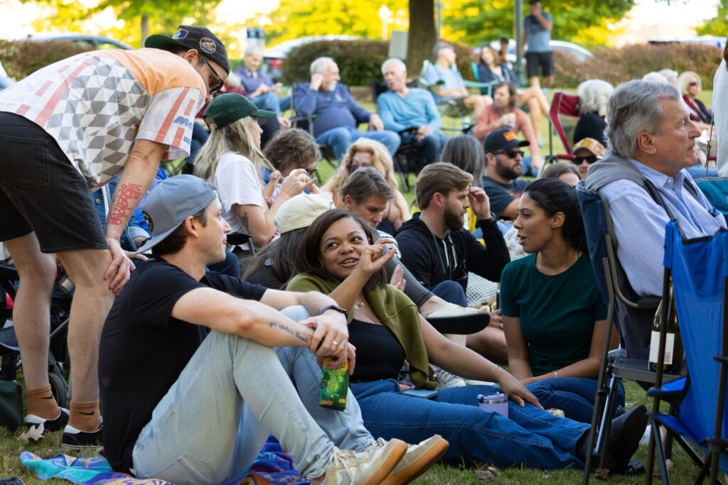 A diverse crowd sits on grass and in some chairs outdoors for a concert.