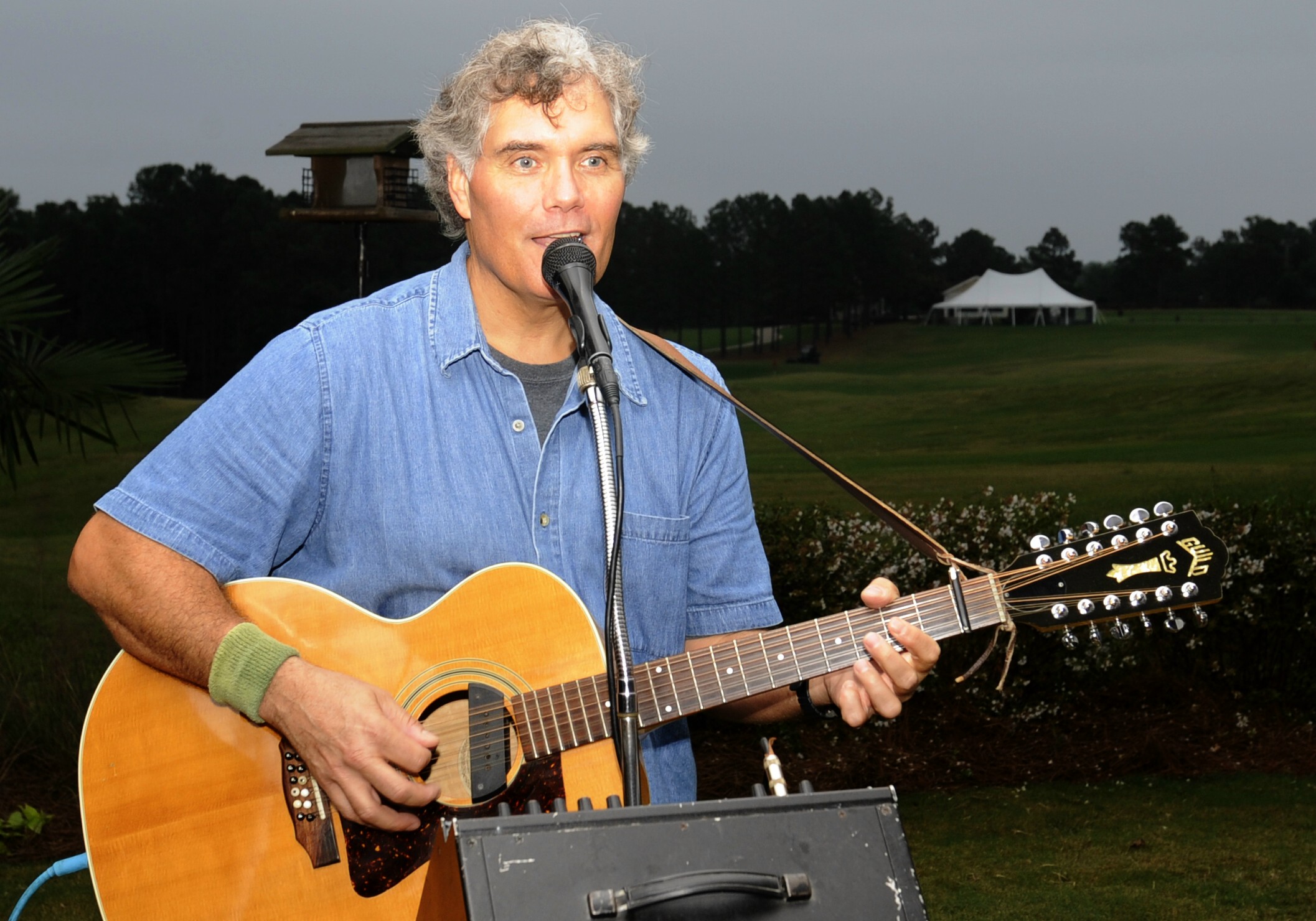 A man with gray hair plays guitar and sings at a microphone at an outdoor venue.