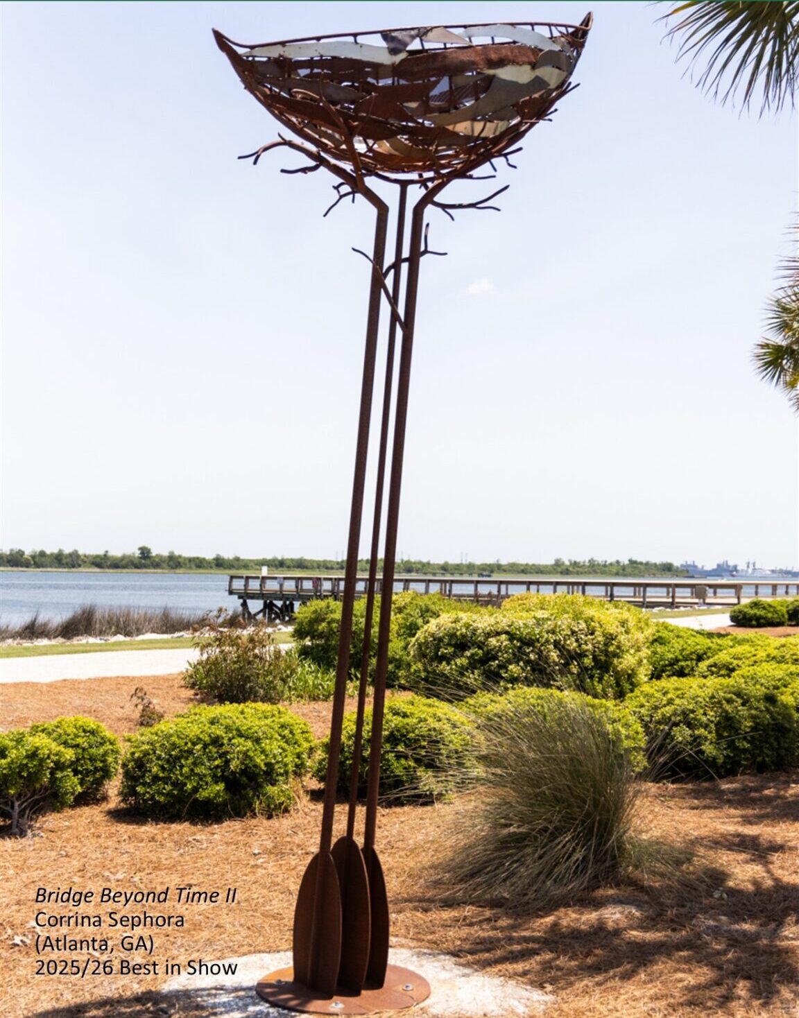 A large outdoor sculpture of brown metal. 3 oars stand on end with branches and a nest at the top.