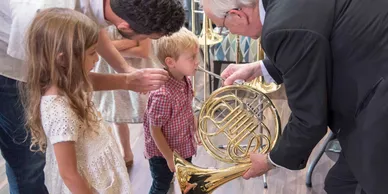 A small boy is introduced to a French horn, as a small girl and 2 men look on.