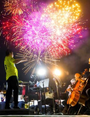 Casually dressed orchestra performs outdoors under a vivid fireworks display.