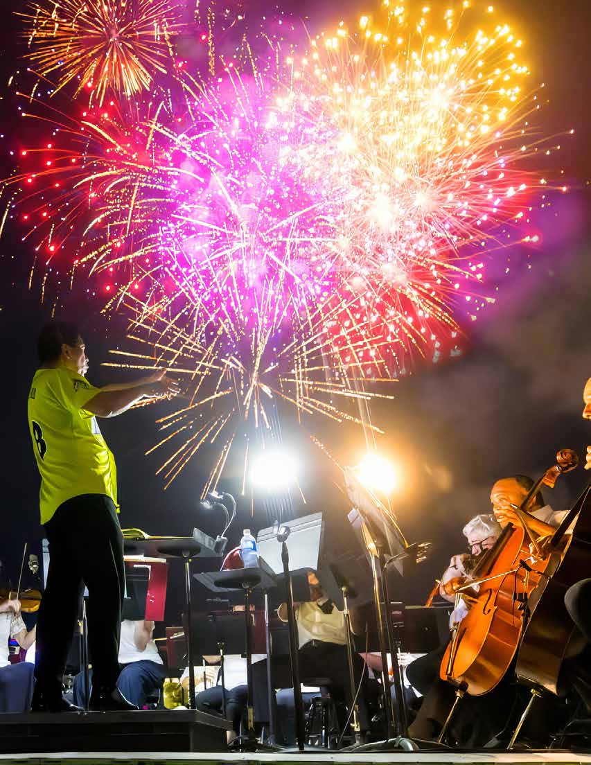 Casually dressed orchestra performs outdoors under a vivid fireworks display.