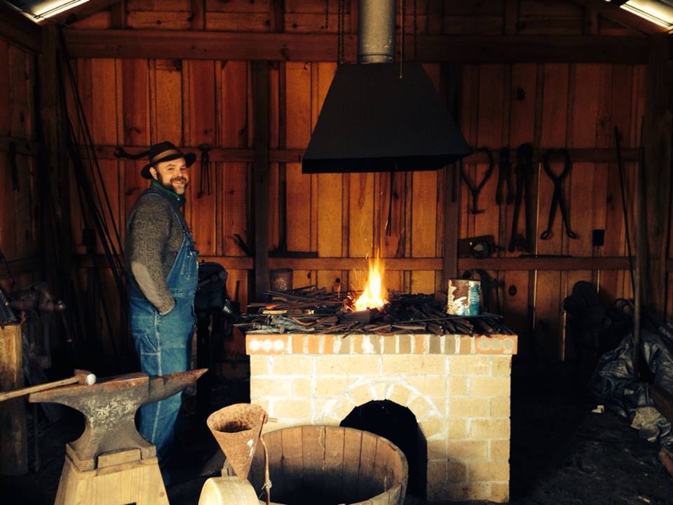 A bearded man smiles as he stands near the open fire in his blacksmithing shop.