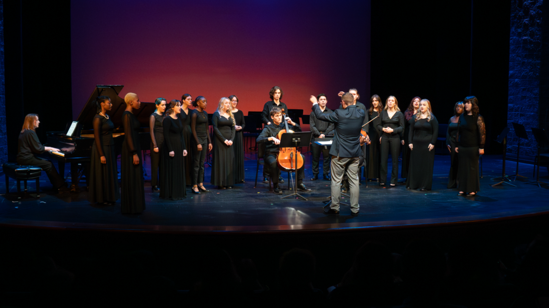A group of student singers perform onstage with cello and piano.