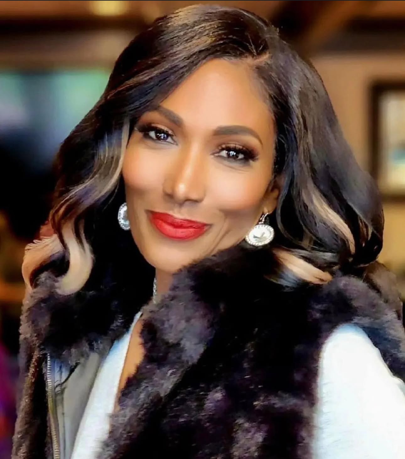 A glamorous Black woman wearing a fur vest over a white shirt smiles at the viewer.