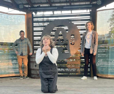 Three actors on an outdoor stage. A woman in overalls kneels in the center, hands to her chin.