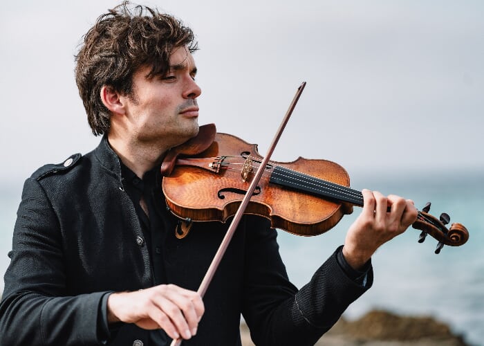 A man with dark hair in a dark suit plays violin near the ocean.