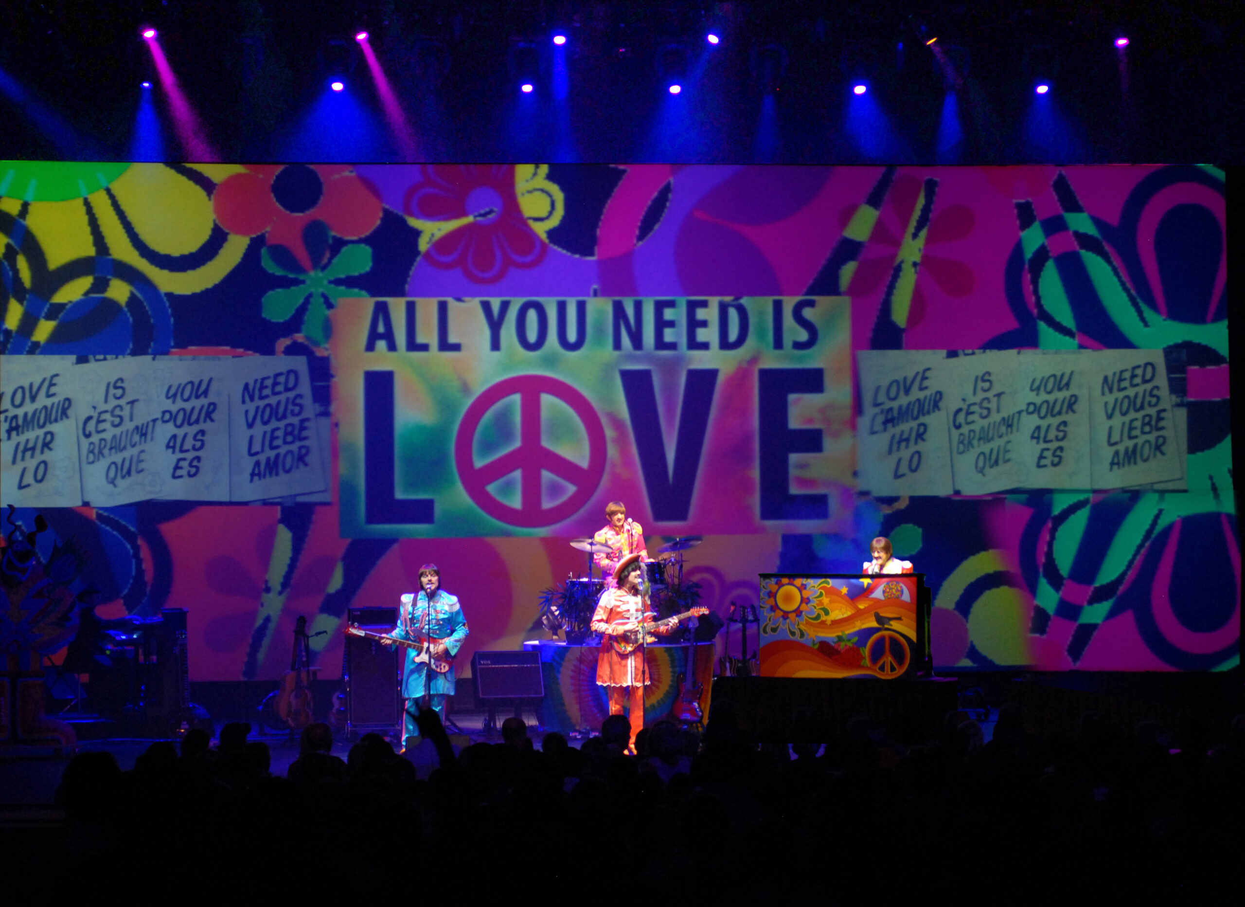 4 costumed men perform as The Beatles on a psychedelic stage. A sign reads, "all you need is love."