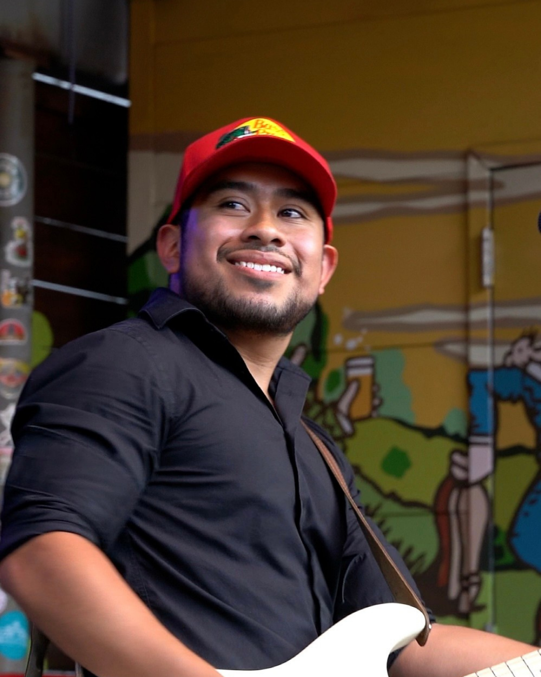 A man presenting as BIPOC smiles as he plays electric guitar against a colorful background.