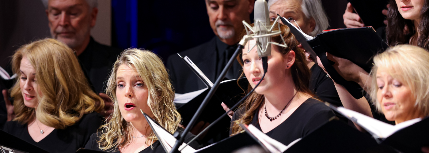 Choral singers dressed in formal black clothing hold folders and perform.