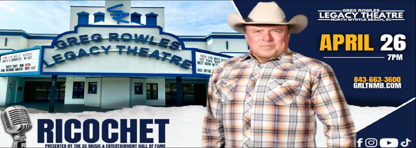 A man in a plaid shirt and cowboy hat poses against an outdoor view of a theatre building.