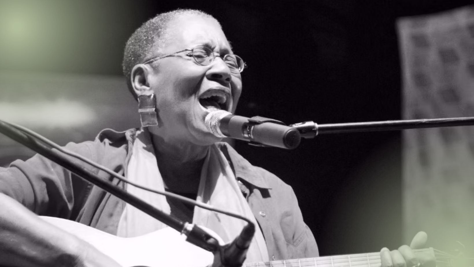 A Black woman with close-cropped hair plays guitar and sings at a microphone.