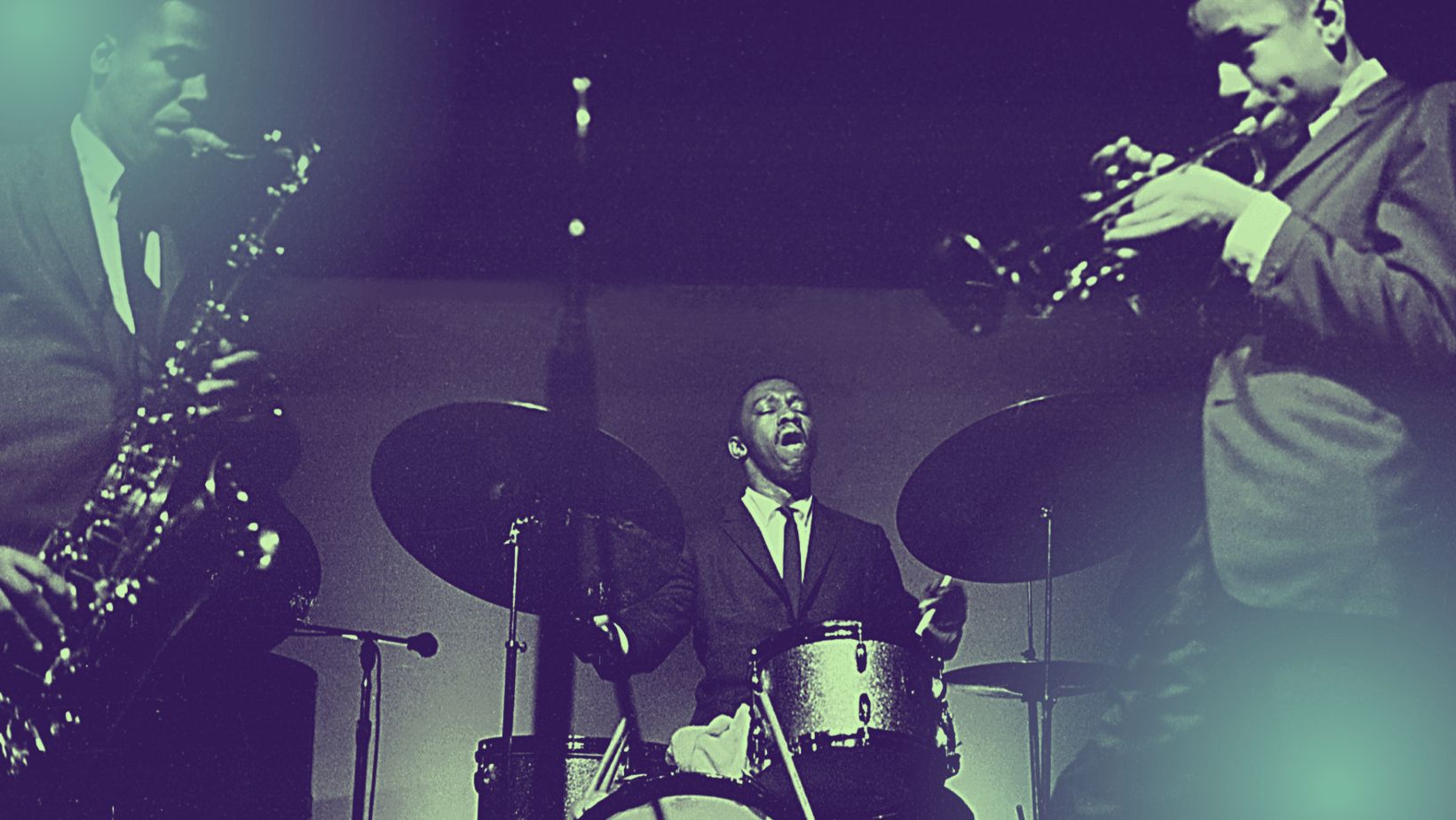 Three Black men in suits perform onstage, playing saxophone, drums, and trumpet.