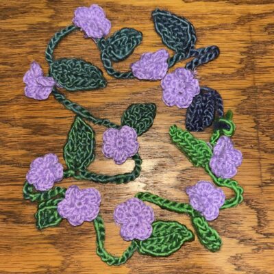 Crocheted purple flowers connected by crocheted green vines and leaves, on a wooden surface.