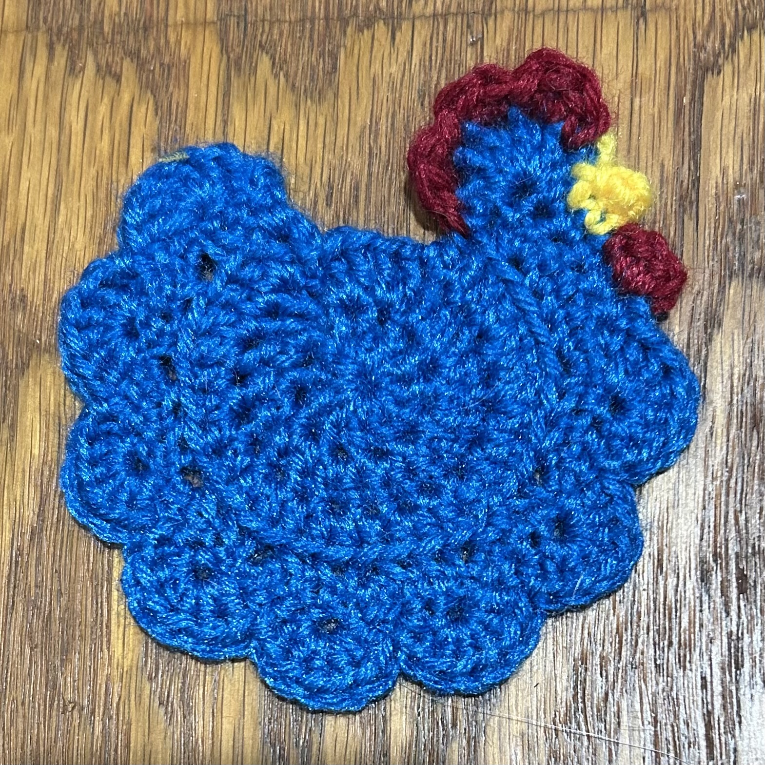 Crochet coaster in the shape of a rooster with a blue circular body, red comb, and yellow beak.