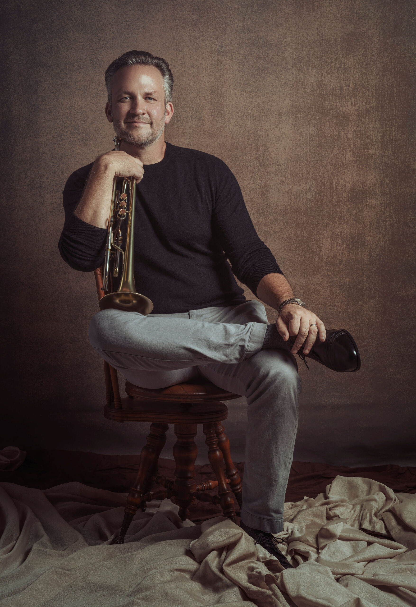 Man with graying hair holding a trumpet sits on a stool with one foot resting on his opposite knee.