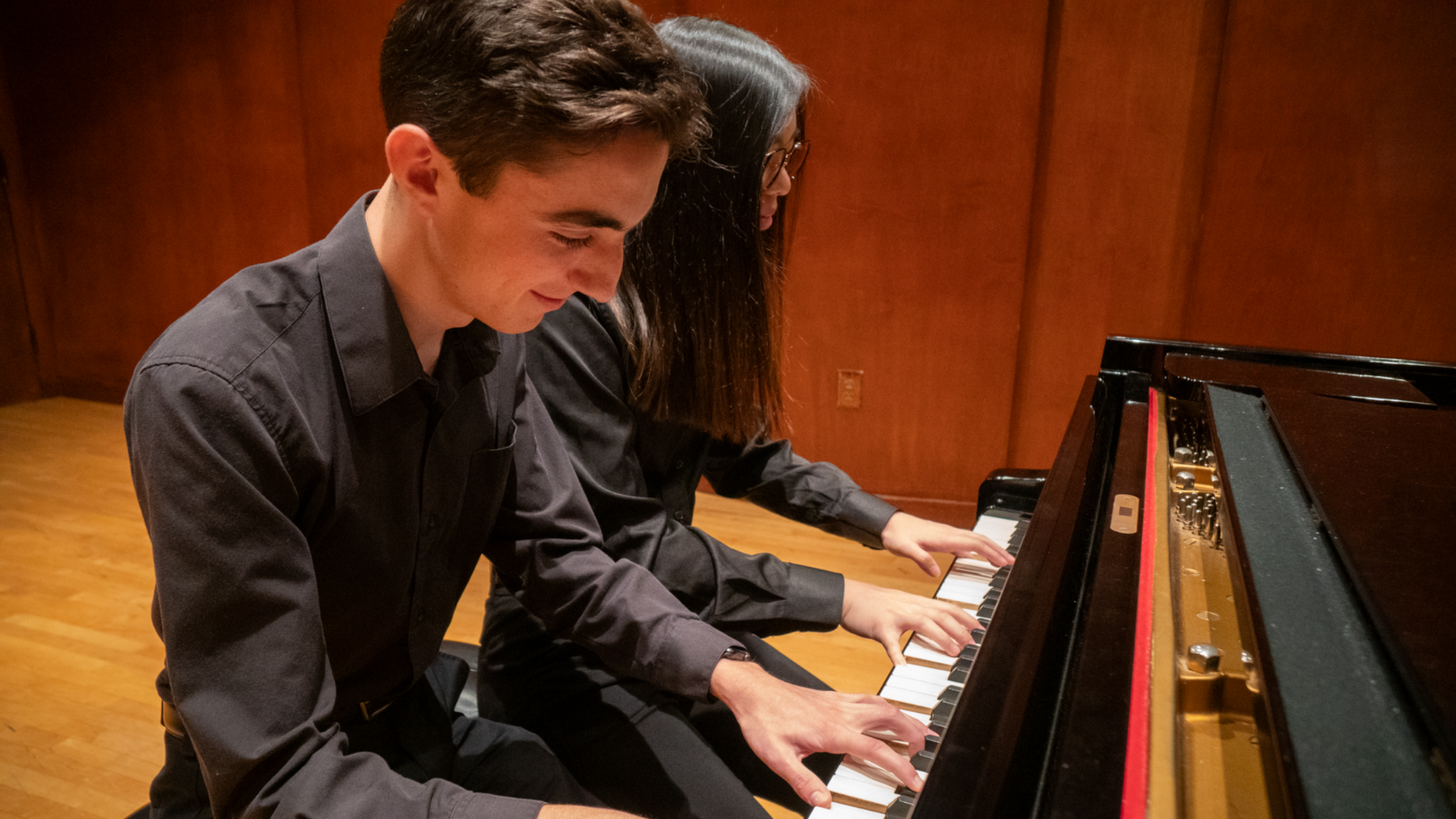 Two students play one piano, dressed in black, on a stage. One has short hair and one has long hair.