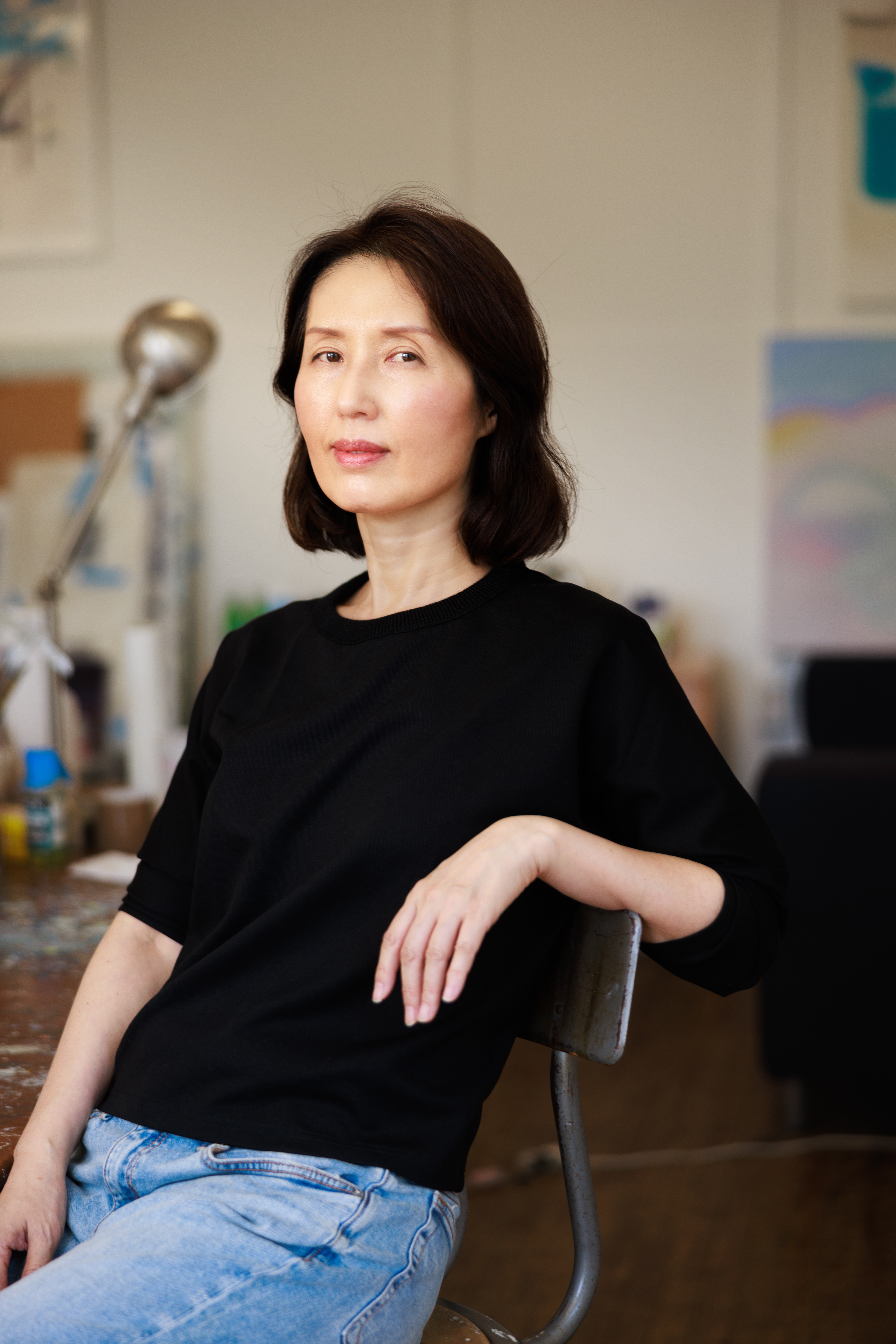 A Korean woman with shoulder-length hair is sitting in a chair in her art studio.