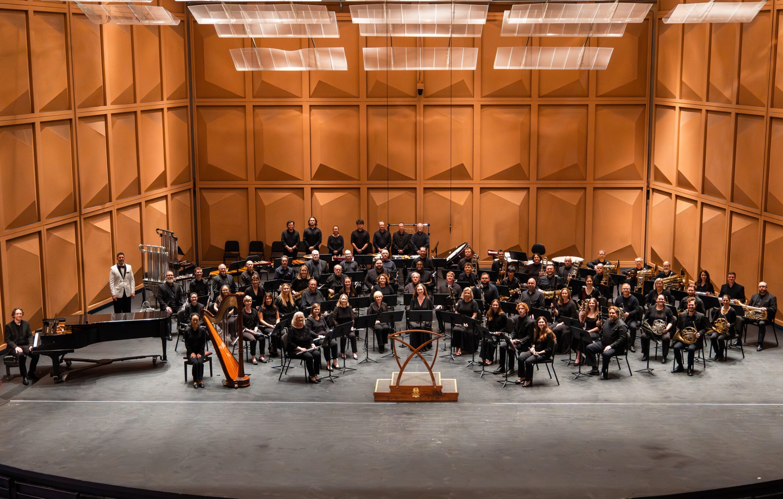 An orchestra of about 80 people sits ready to perform on a large stage.