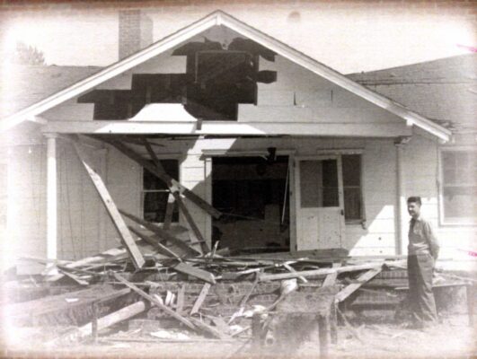 A man stands outside a badly damaged house with lumber and debris.