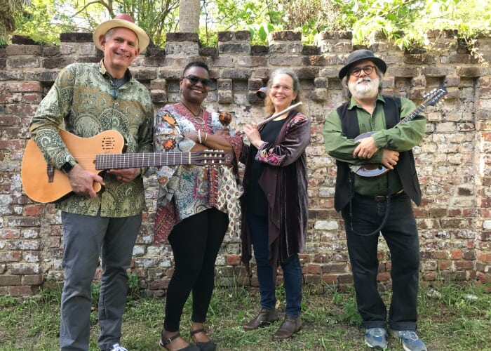 Four smiling musicians pose outdoors by a brick wall, with stringed instruments and a flute.